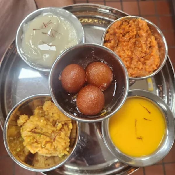A silver platter with five bowls of Indian desserts, including gulab jamun, halwa, and kheer.
