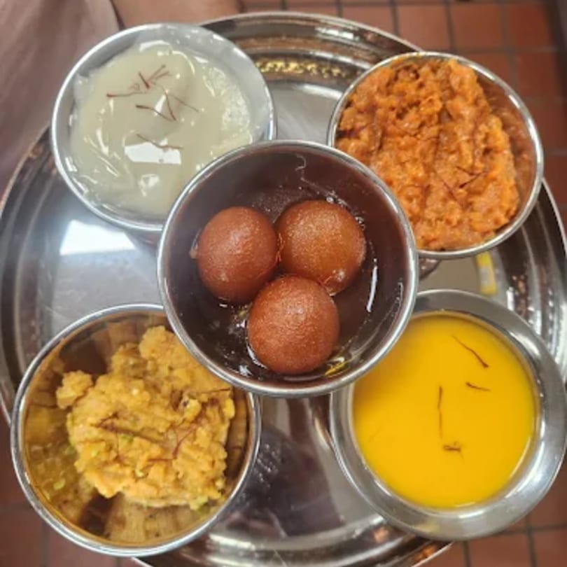 A silver platter with five bowls of Indian desserts, including gulab jamun, halwa, and kheer.