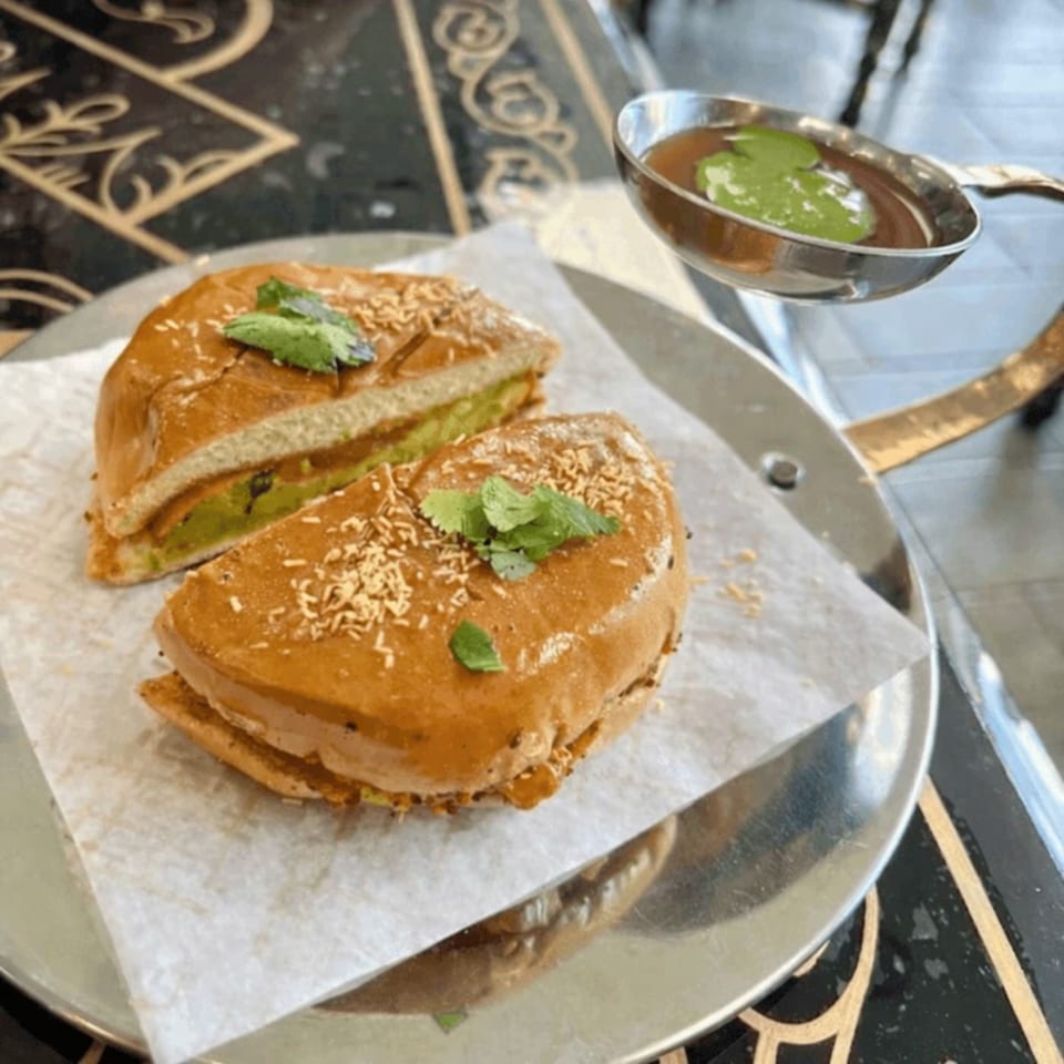Indian street food sandwich, cut in half, with a side of dipping sauce in a small metal bowl.