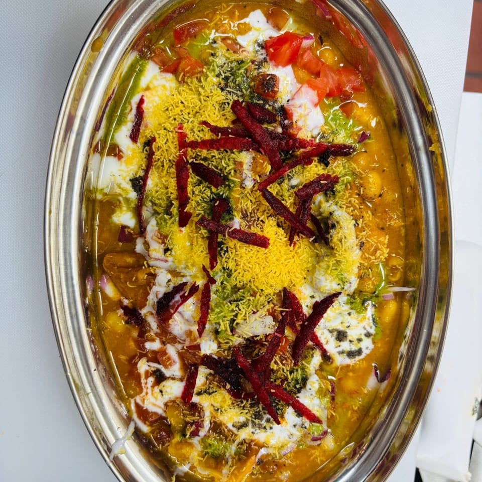 Close-up of a vibrant Indian street food dish, MaanKi Thali, with sauces, toppings, and red garnish.