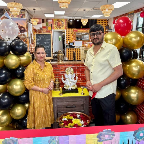 Two people standing in front of a Ganesha statue, balloons, and Indian restaurant decor.