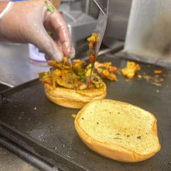 A gloved hand uses a spatula to add Indian street food filling to a toasted bun on a griddle.