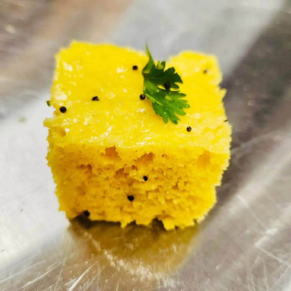 A square piece of yellow Dhokla, garnished with mustard seeds and a sprig of cilantro.