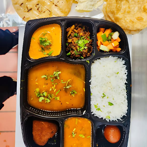 A MaanKi Thali Indian meal with rice, curries, vegetables, and dessert in a black partitioned container.