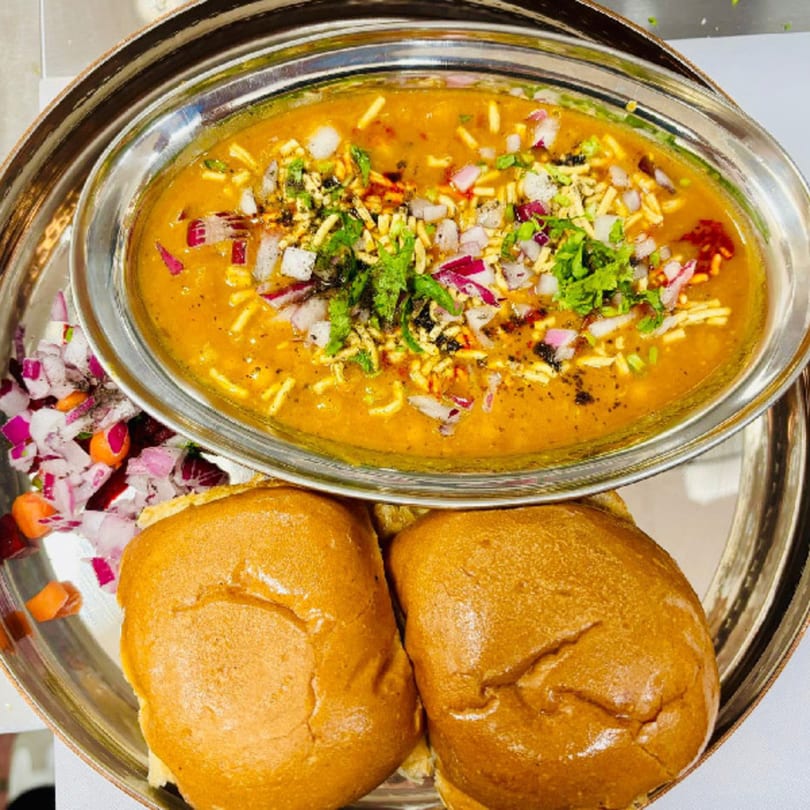 MaanKi Thali's Misal Pav, a spicy Indian curry with pav bread, onions, and cilantro.